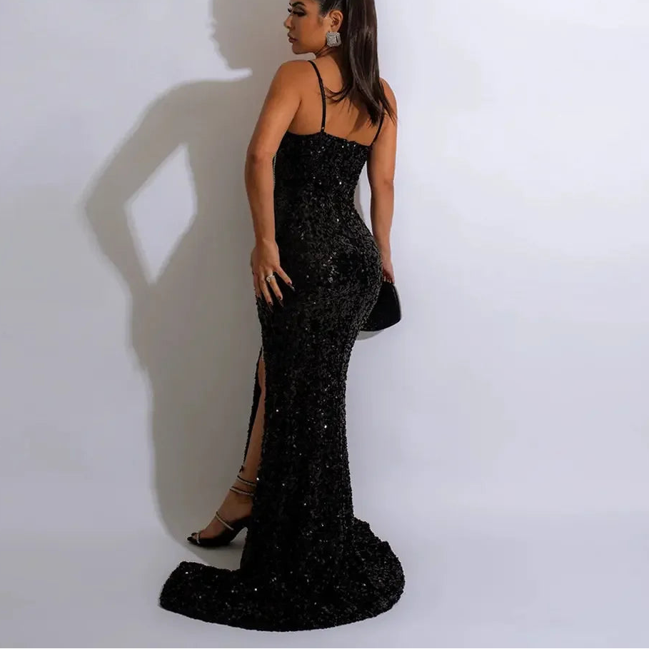 Woman in a black sequin dress with a high slit, standing against a white wall.